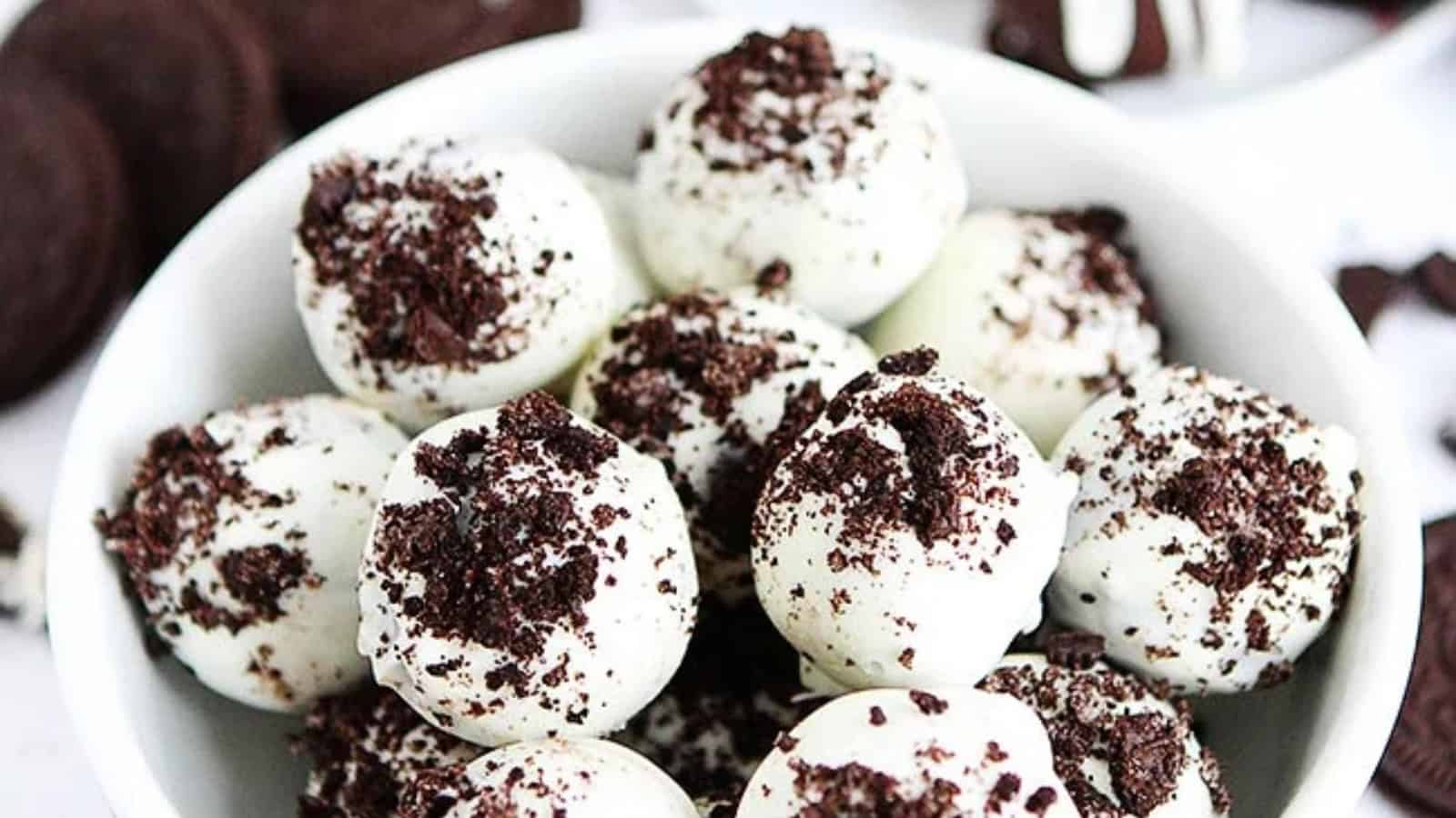 A bowl of white chocolate truffles topped with crumbled chocolate cookies. The truffles are arranged closely together, creating an appetizing display of contrasting colors and textures.