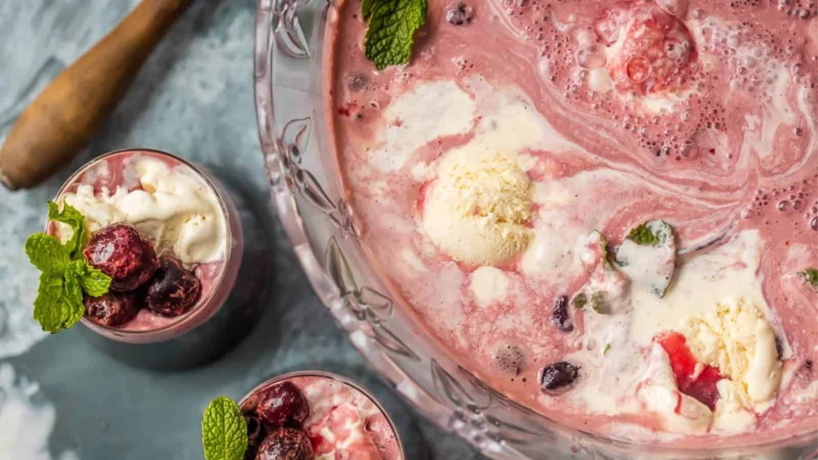 A large glass bowl filled with a creamy pink punch containing scoops of ice cream and mixed berries. Two glasses beside it also have the punch, garnished with mint leaves and berries. A wooden spoon is partially visible on the side.