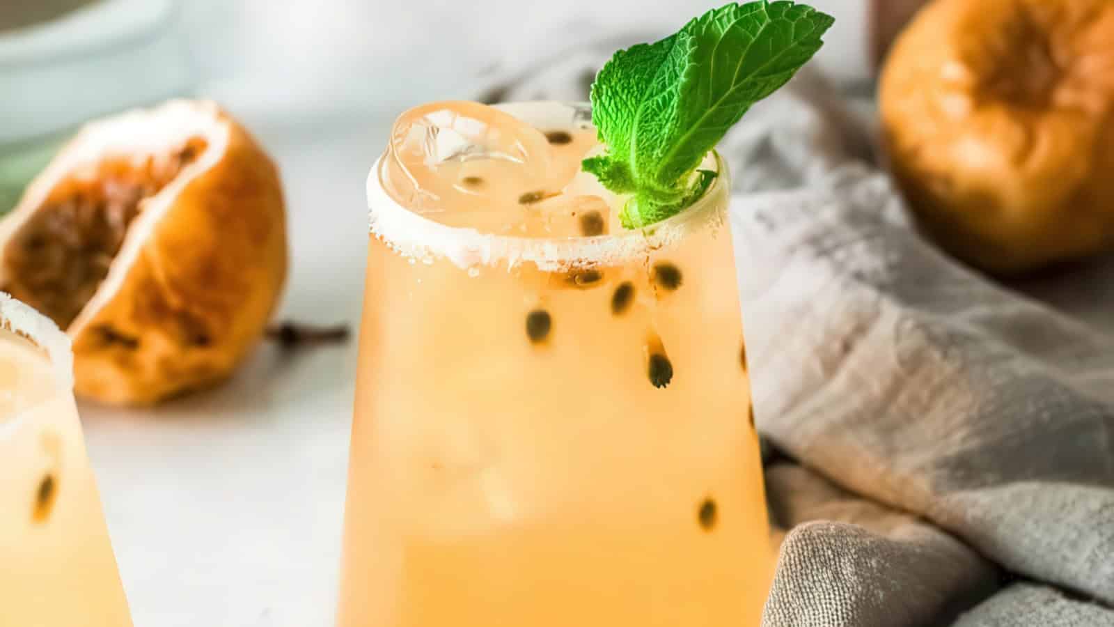 A refreshing glass of passion fruit juice with ice, garnished with a sprig of mint. In the background, there are passion fruits and a light gray cloth. The drink is in a clear glass on a white surface.
