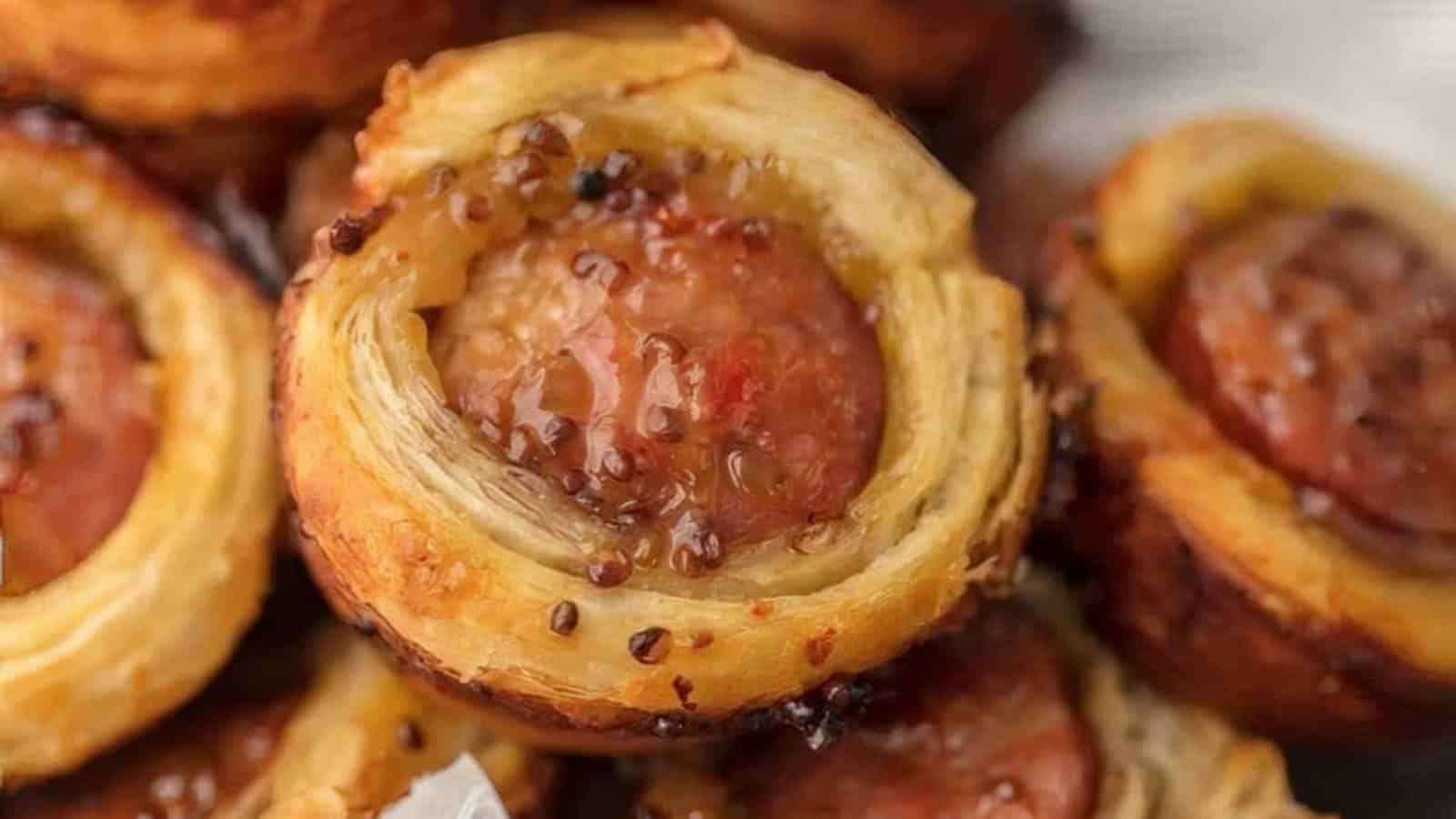 Close-up of a savory pastry showing a golden-brown puff pastry wrapped around a sausage, topped with a glossy layer of mustard or sauce. The pastry looks crispy and appetizing, with seeds visible in the topping.