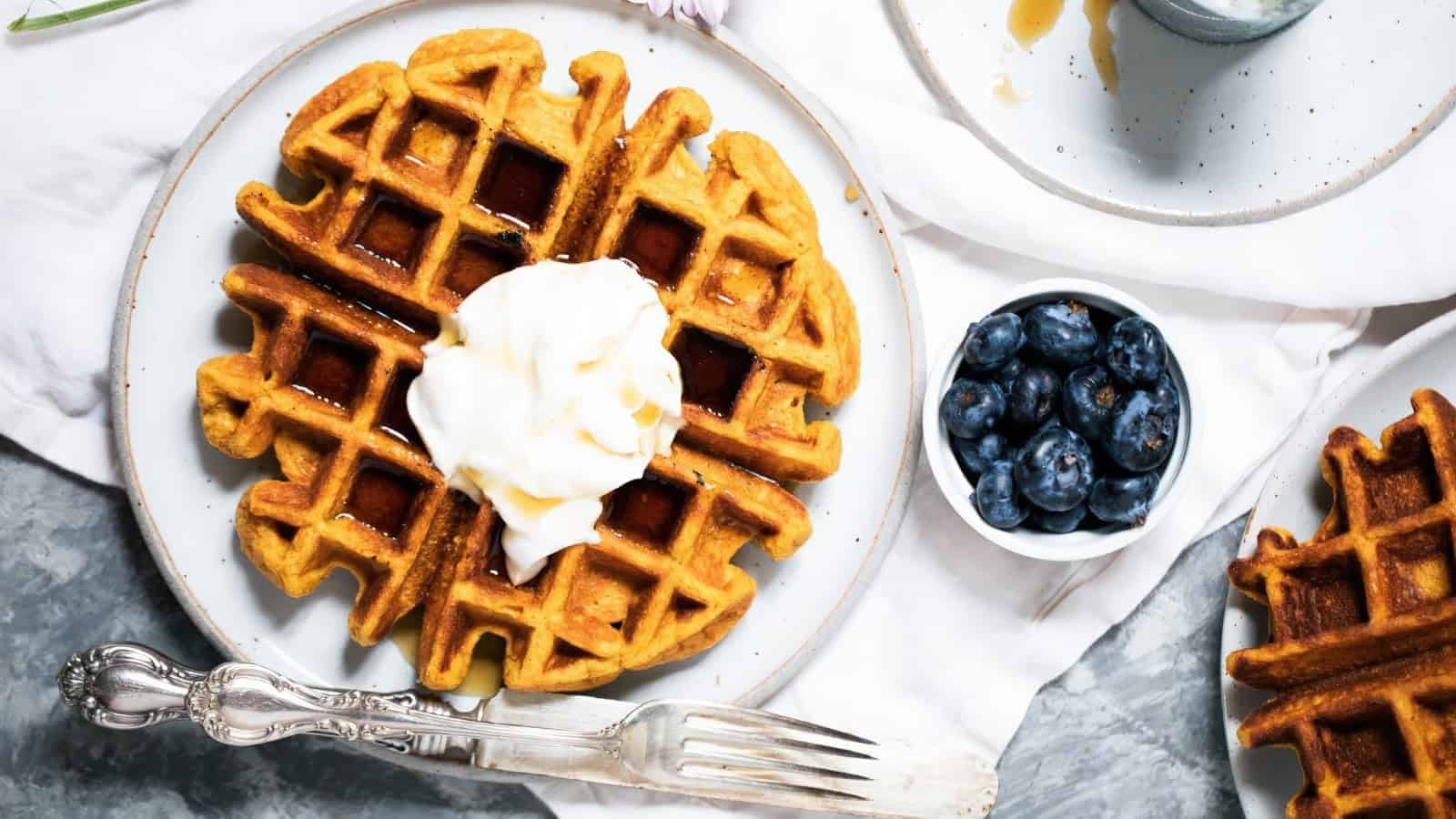 A round waffle with whipped cream and syrup on a white plate, accompanied by a small bowl of blueberries. A fork and knife are placed beside the plate on a light-colored napkin.
