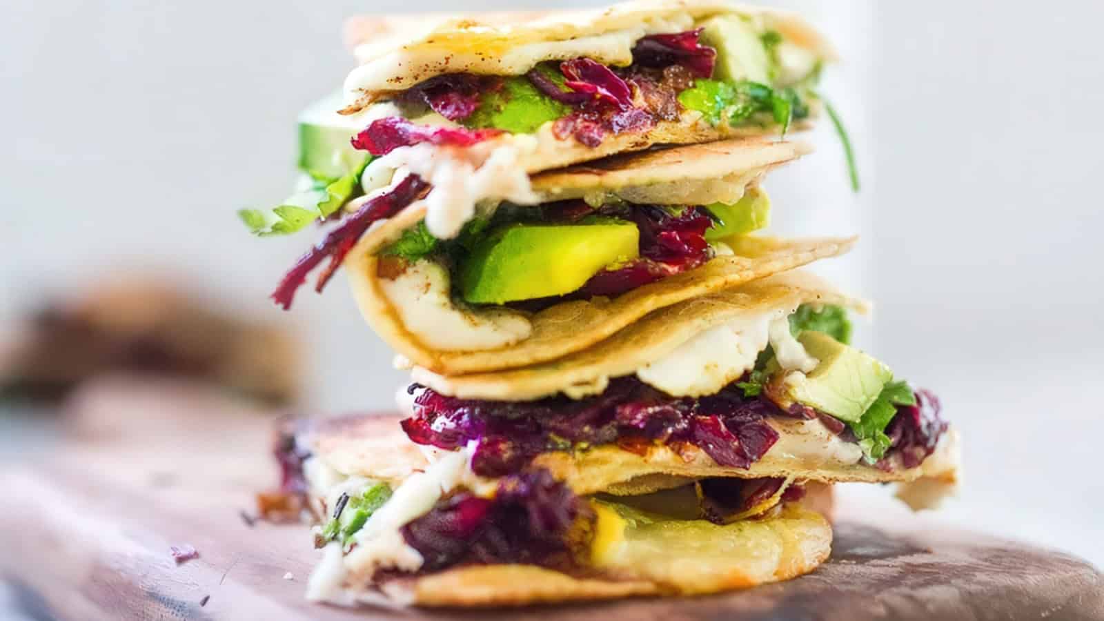 A stack of freshly made quesadillas filled with avocado, melted cheese, greens, and vibrant red cabbage, placed on a wooden cutting board. The layers are visible, showcasing the colorful ingredients.