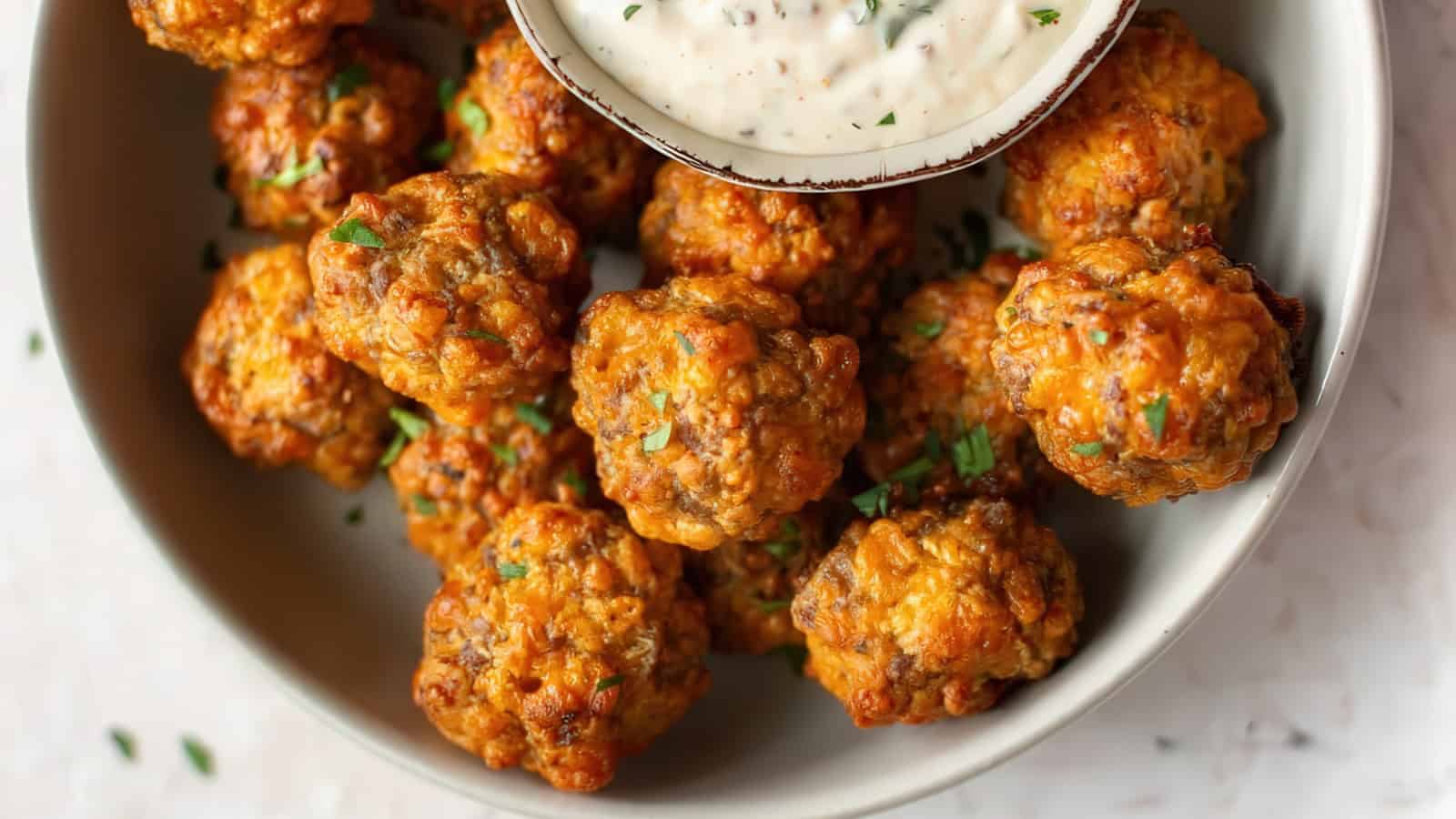A plate of golden-brown sausage balls garnished with herbs, surrounding a small bowl of white dipping sauce. The texture of the sausage balls looks crispy and savory.