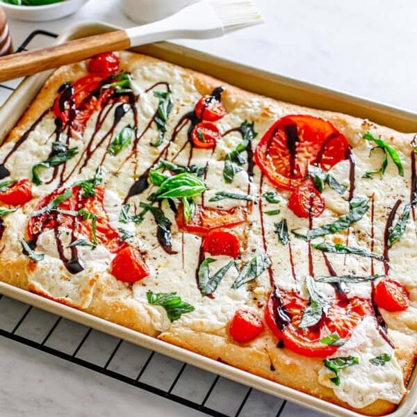 Rectangular focaccia bread topped with mozzarella, sliced tomatoes, fresh basil, and a drizzle of balsamic glaze on a wire rack.