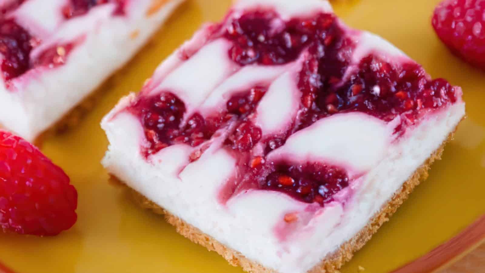 A close-up of a slice of raspberry swirl cheesecake on a yellow plate. The dessert has a graham cracker crust, creamy white filling with swirls of raspberry topping. Fresh raspberries are placed nearby.