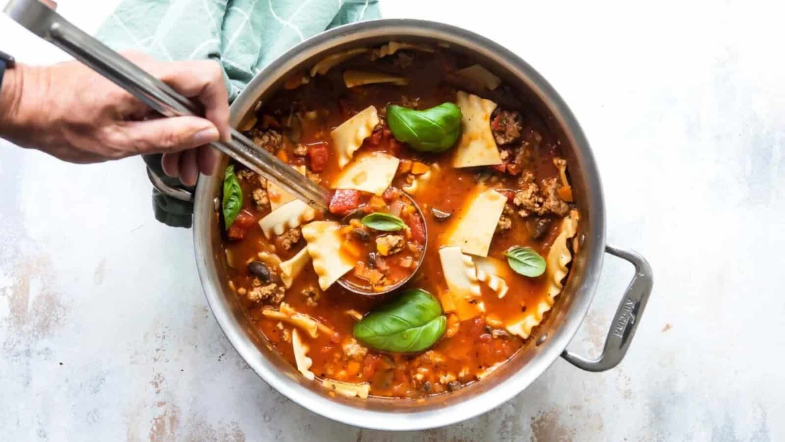 A person holds a ladle over a pot of lasagna soup, filled with tomatoes, pasta pieces, minced meat, and basil leaves. The pot rests on a light surface with a green cloth nearby.