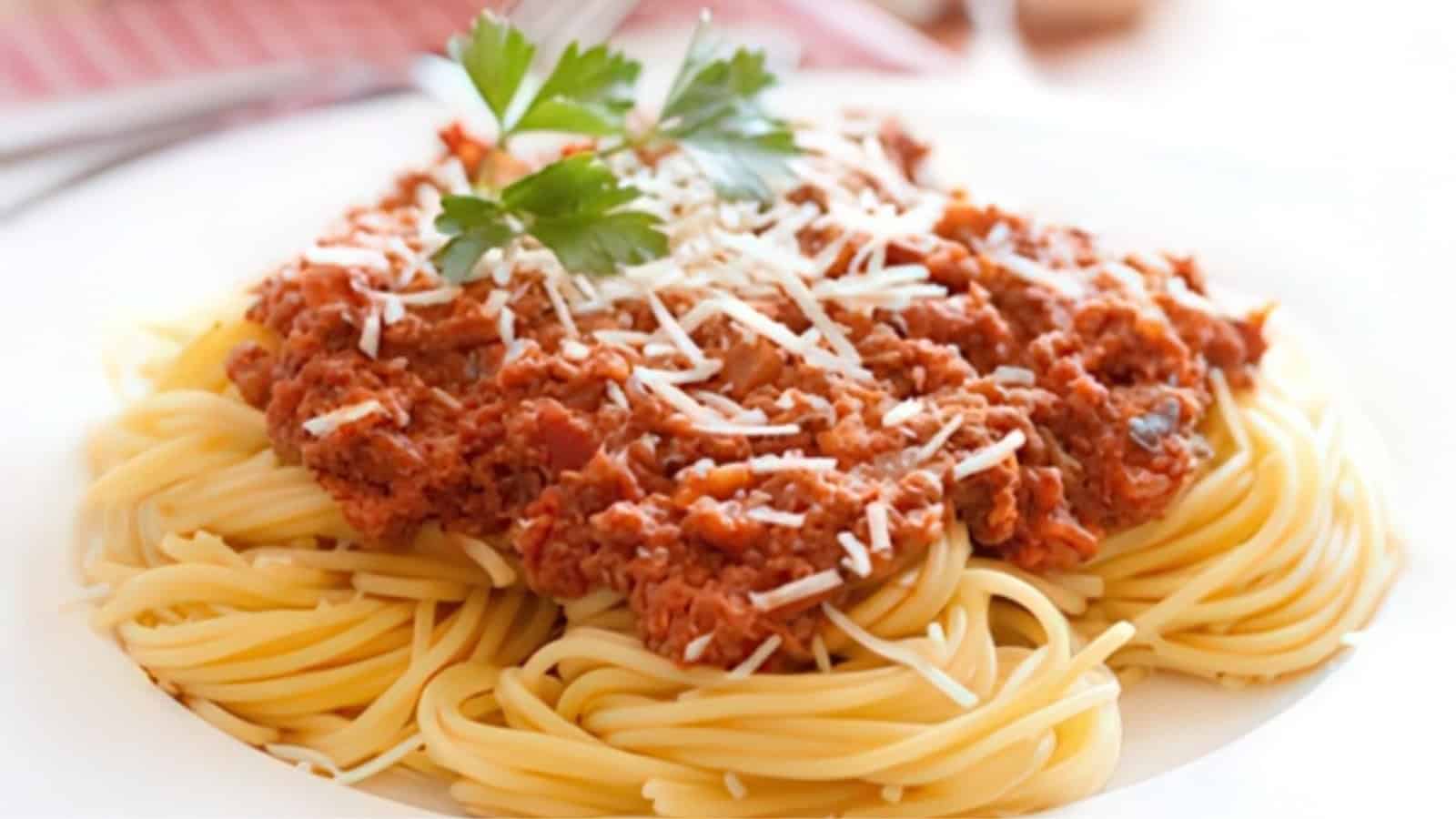 A plate of spaghetti topped with tomato-based meat sauce and garnished with grated cheese and fresh parsley. The dish is served on a white plate, creating a colorful contrast with the red sauce and green parsley.