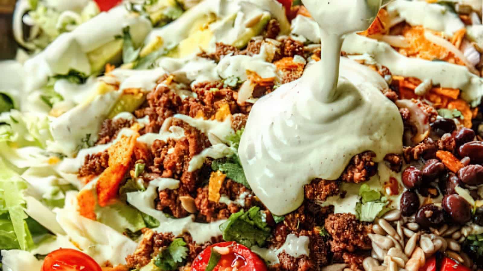 Close-up of a vibrant taco salad featuring seasoned ground meat, cherry tomatoes, black beans, shredded cheese, and avocado, topped with a drizzle of creamy dressing. Bright and fresh presentation with a variety of textures and colors.