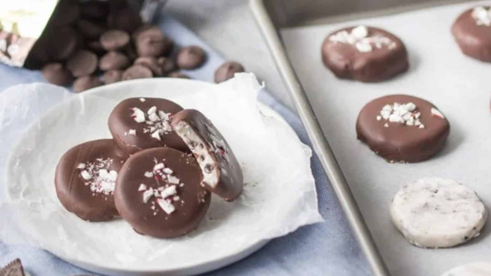 The image shows chocolate-covered cookies on a plate and a baking sheet. Some cookies are topped with crushed peppermint, and one cookie has a bite taken out of it, revealing a cream filling. Chocolate chips are scattered in the background.
