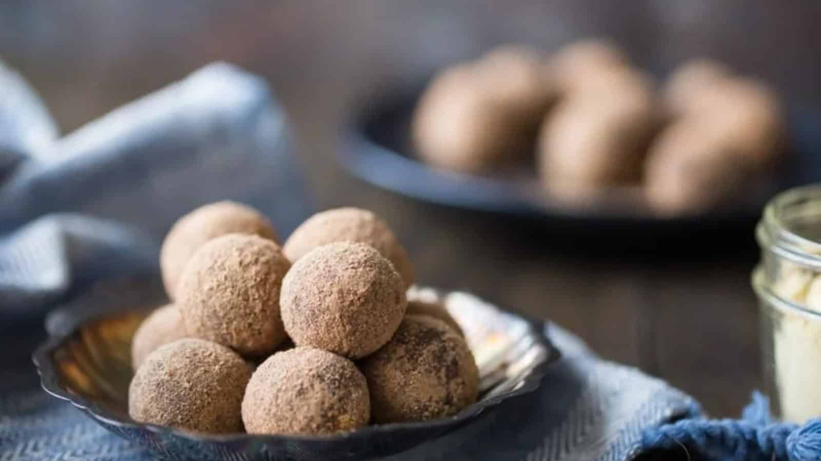 A close-up of a bowl filled with cocoa-dusted truffles arranged in a pyramid shape. The bowl is placed on a wooden surface with a blue cloth beside it. In the background, another plate with similar truffles is visible, slightly out of focus.