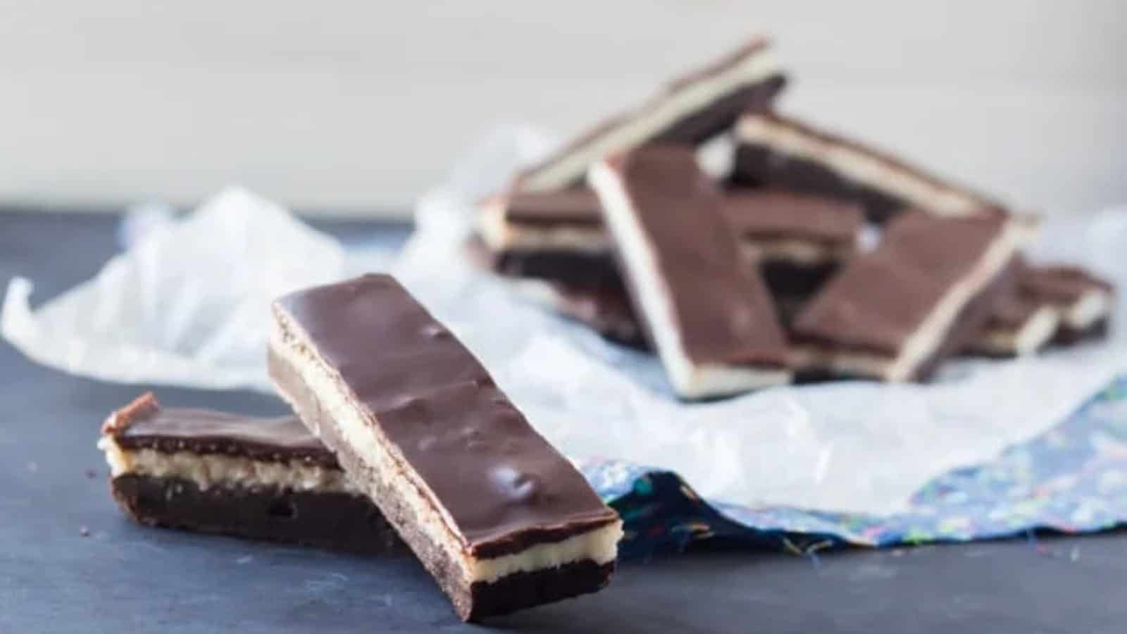 Chocolate bars with layered cream filling are arranged on a dark surface. Some bars are neatly stacked on crinkled paper, while two are placed in the foreground. The background is softly blurred, highlighting the rich texture of the bars.