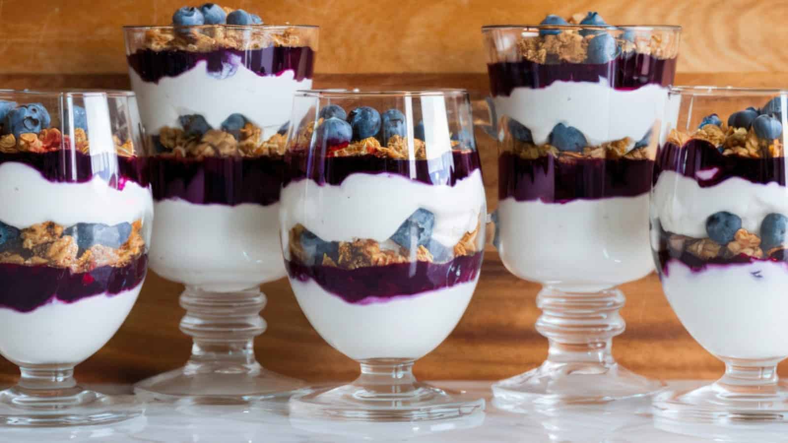 Five clear glasses filled with layers of yogurt, blueberry sauce, granola, and fresh blueberries, arranged on a wooden background. The vibrant colors and textures create an appealing dessert display.