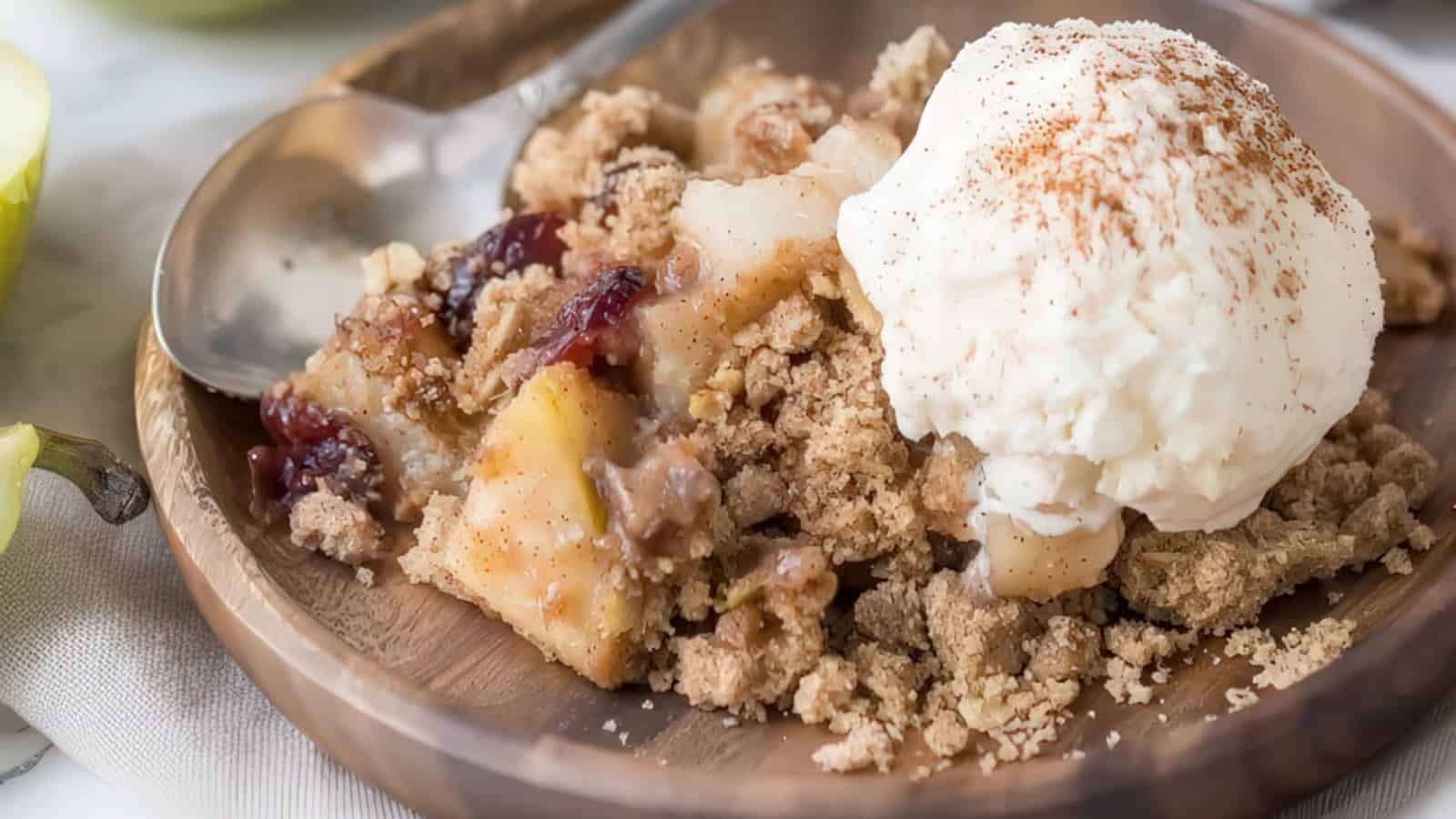 A wooden plate holds a serving of apple crumble topped with a scoop of vanilla ice cream and a sprinkle of cinnamon. A spoon rests beside the dessert, ready for use. The crumble has visible pieces of apple and hints of red berries.
