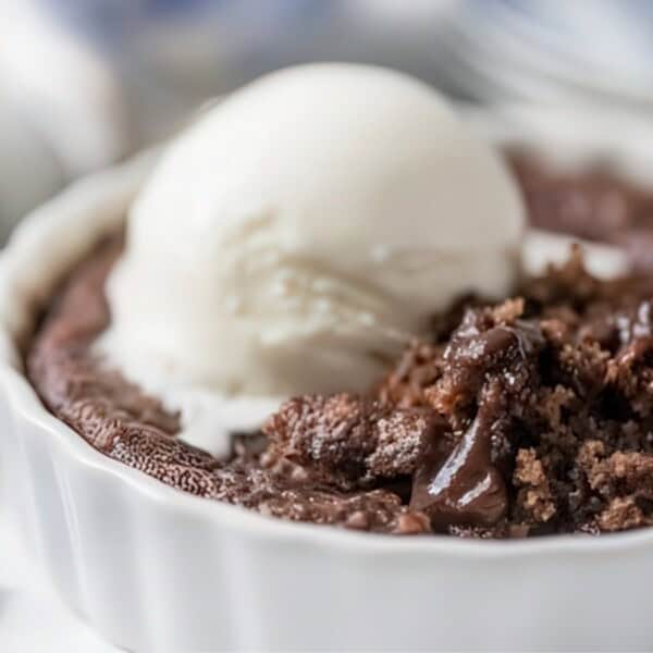 A close-up of a chocolate lava cake in a white ramekin topped with a scoop of vanilla ice cream. A spoon is partially visible, digging into the soft, gooey cake. The surface is slightly cracked, revealing its rich texture.