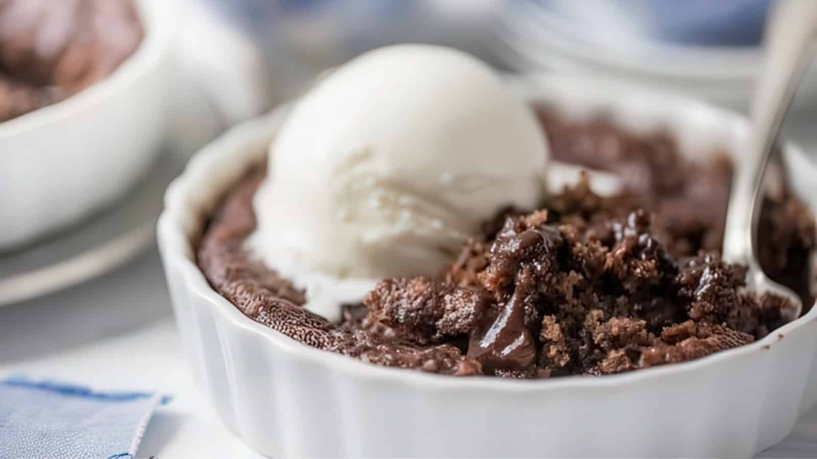 A close-up of a chocolate lava cake in a white ramekin topped with a scoop of vanilla ice cream. A spoon is partially visible, digging into the soft, gooey cake. The surface is slightly cracked, revealing its rich texture.