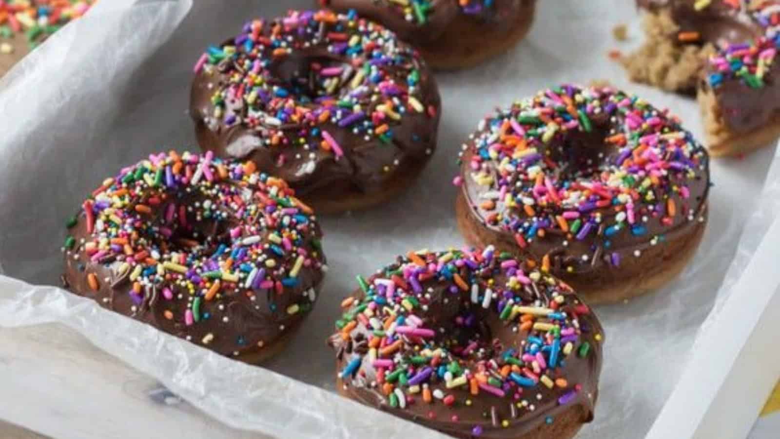 A tray of chocolate-glazed doughnuts topped with colorful sprinkles, placed on wax paper.