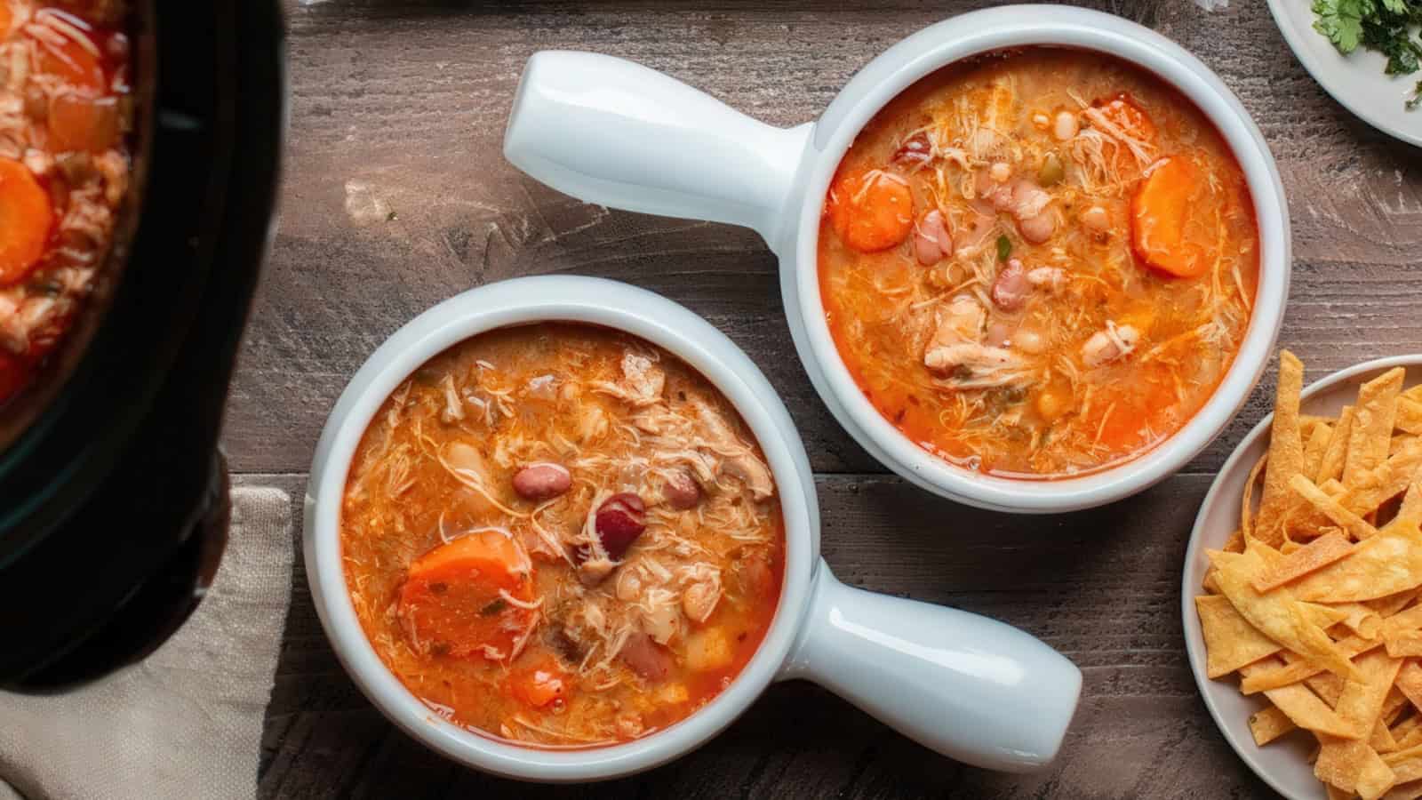 Two white bowls filled with hearty chicken soup, featuring shredded chicken, beans, and carrot slices, are placed on a wooden table. Beside them is a plate of crispy tortilla strips.