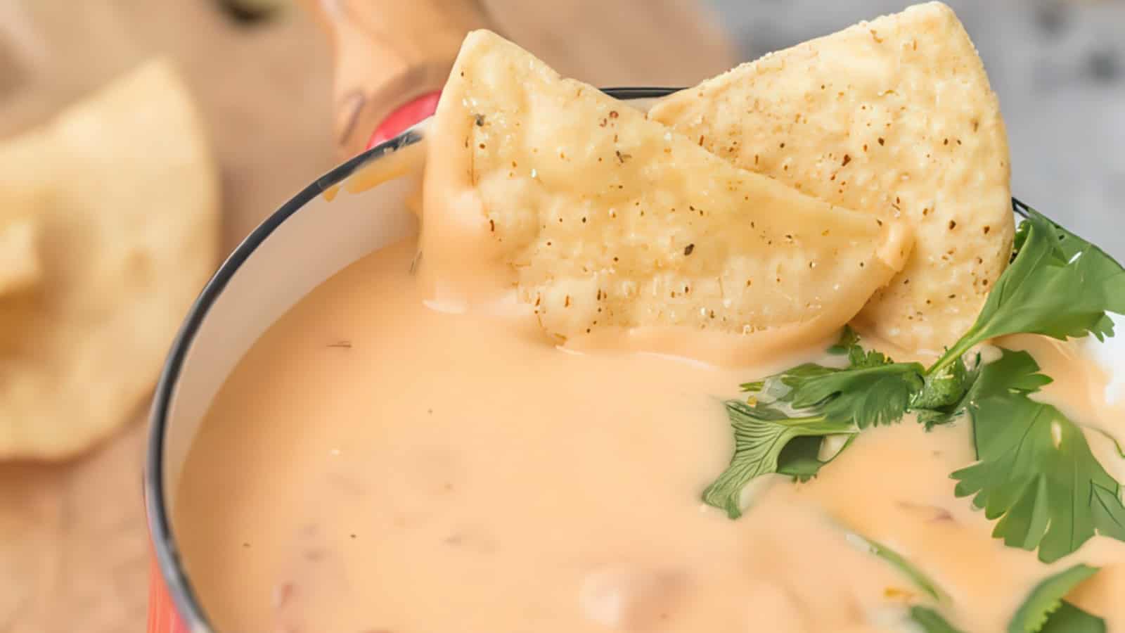 A bowl of creamy cheese dip with two tortilla chips dipped in it. Fresh cilantro leaves garnish the dip. A wooden board with more chips is partially visible in the background.