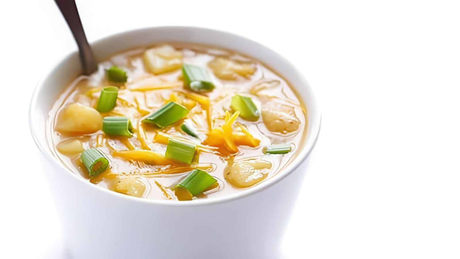 A bowl of creamy potato soup garnished with shredded cheese and chopped green onions, with a spoon on the side. The soup appears warm and comforting, with visible chunks of potatoes.