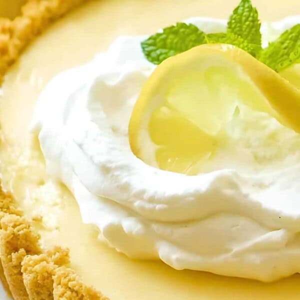 A lemon pie topped with whipped cream, a lemon slice, and mint garnish. The crust is crumbly with a smooth filling.