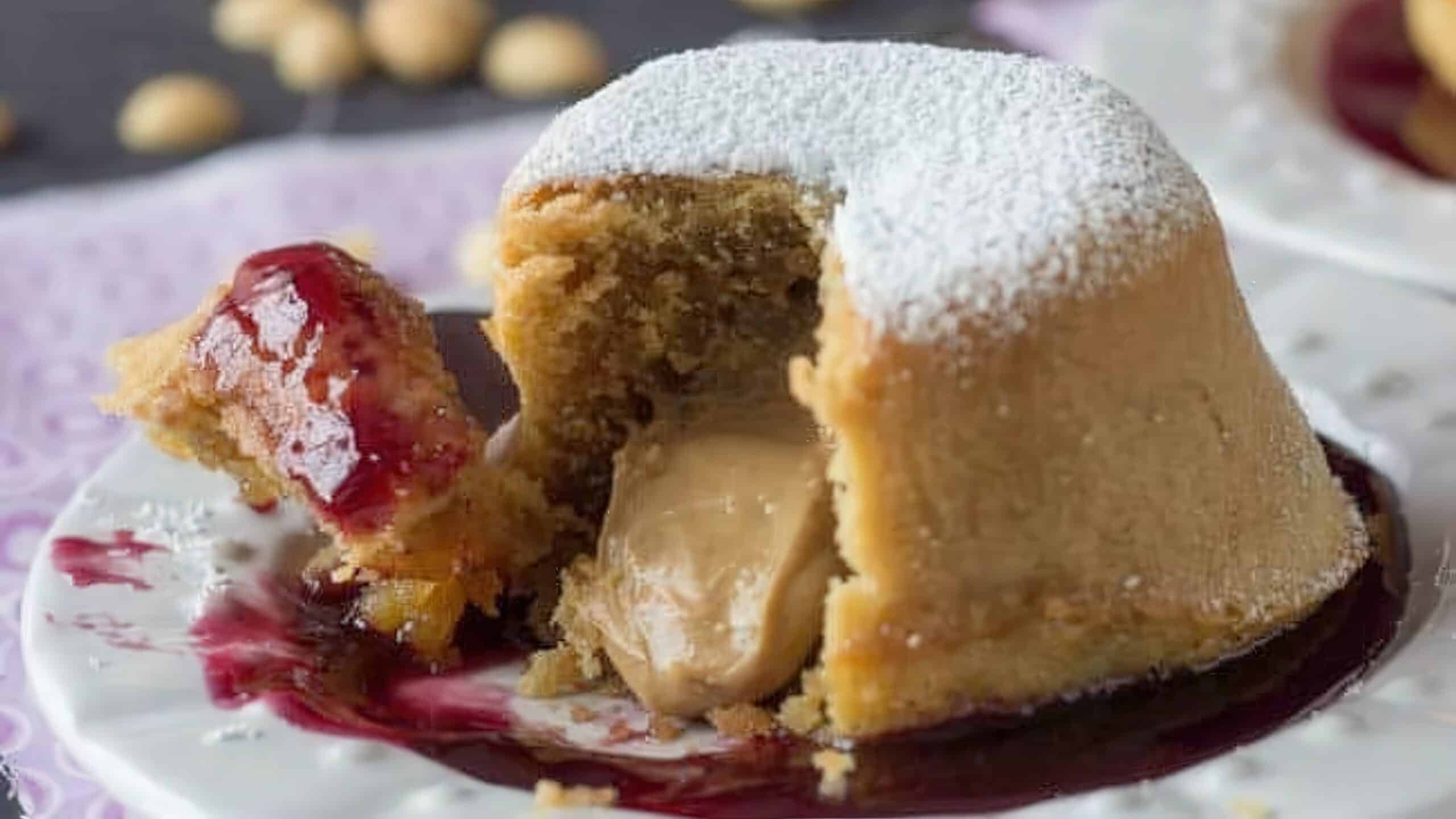 A sliced dessert reveals a creamy caramel filling inside a sponge cake, dusted with powdered sugar. It sits on a white plate with a drizzle of berry sauce on one side.