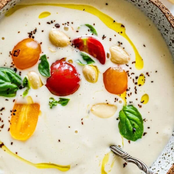A bowl of creamy soup garnished with colorful cherry tomatoes, almonds, basil leaves, and a drizzle of olive oil. A spoon rests in the bowl, and the dish is presented in a rustic, speckled bowl.