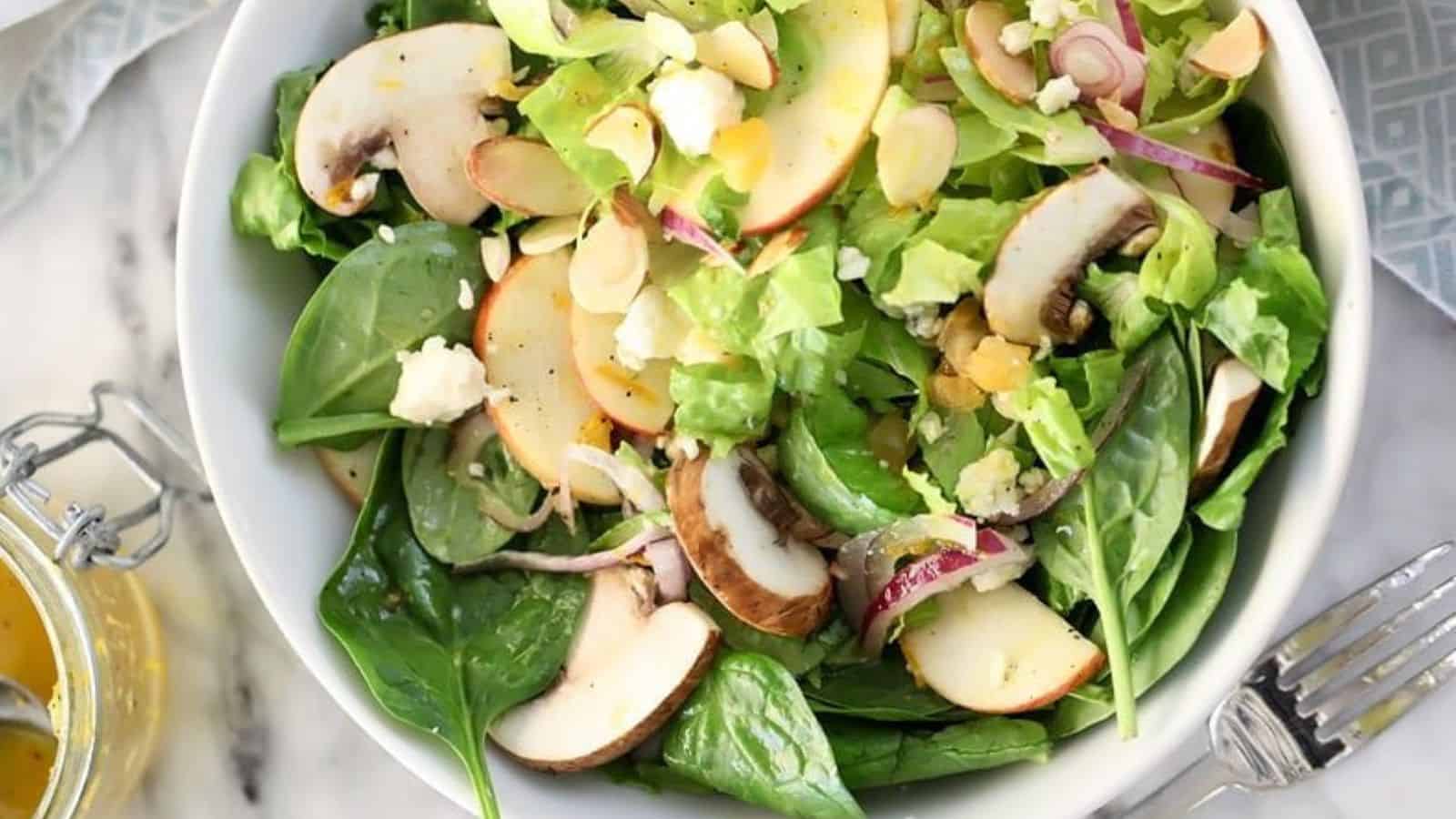 A fresh salad in a white bowl featuring spinach leaves, apple slices, mushrooms, red onion, and lettuce, garnished with almonds and crumbled cheese. A jar of dressing is nearby, and a fork rests beside the bowl.