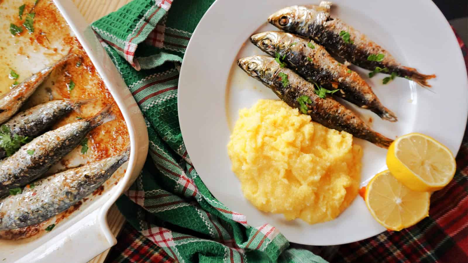 A plate of four grilled sardines garnished with herbs, served alongside a portion of creamy polenta and two lemon halves. The background includes a checkered cloth and a baking dish with additional sardines.