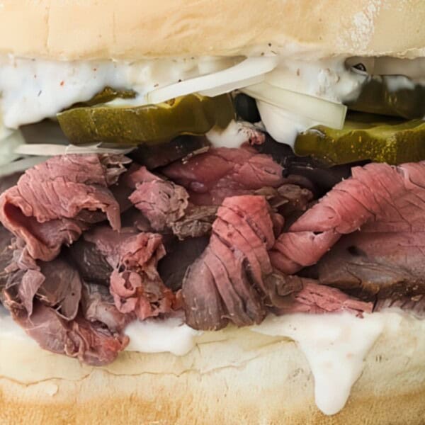 Close-up of a roast beef sandwich stacked on a soft bun. Layers include thinly sliced roast beef, creamy sauce, pickle slices, and shreds of onion. The filling slightly overflows, showcasing a hearty and indulgent meal.