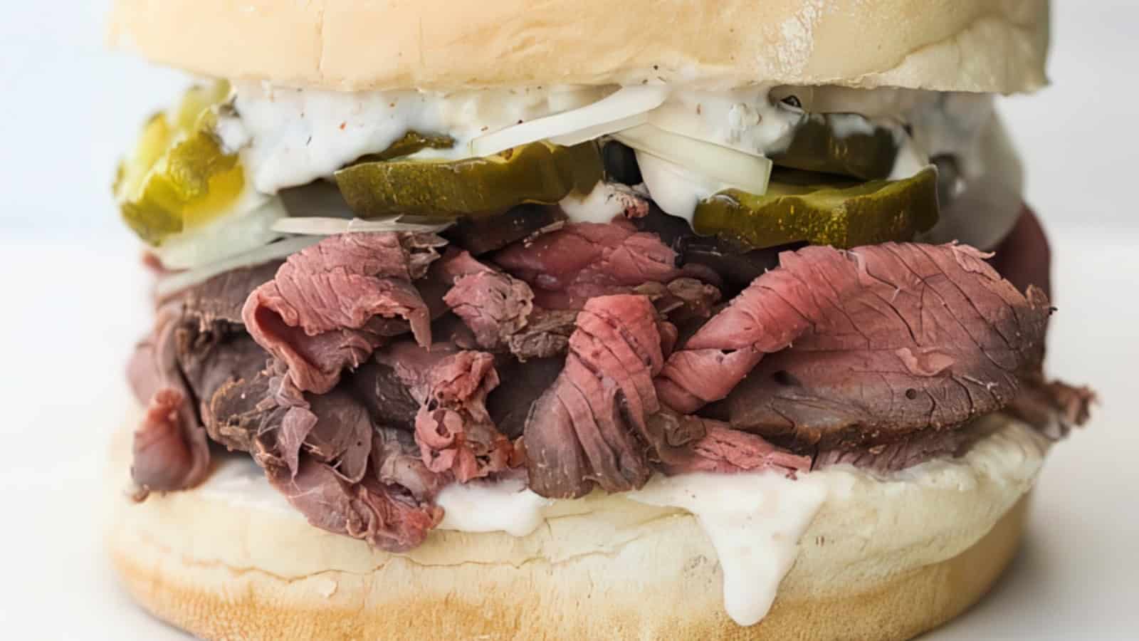 Close-up of a roast beef sandwich stacked on a soft bun. Layers include thinly sliced roast beef, creamy sauce, pickle slices, and shreds of onion. The filling slightly overflows, showcasing a hearty and indulgent meal.