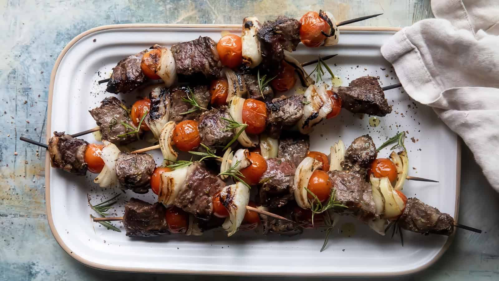 A platter of grilled beef skewers with cherry tomatoes and onion slices, garnished with fresh rosemary sprigs. A white napkin is placed beside the dish.
