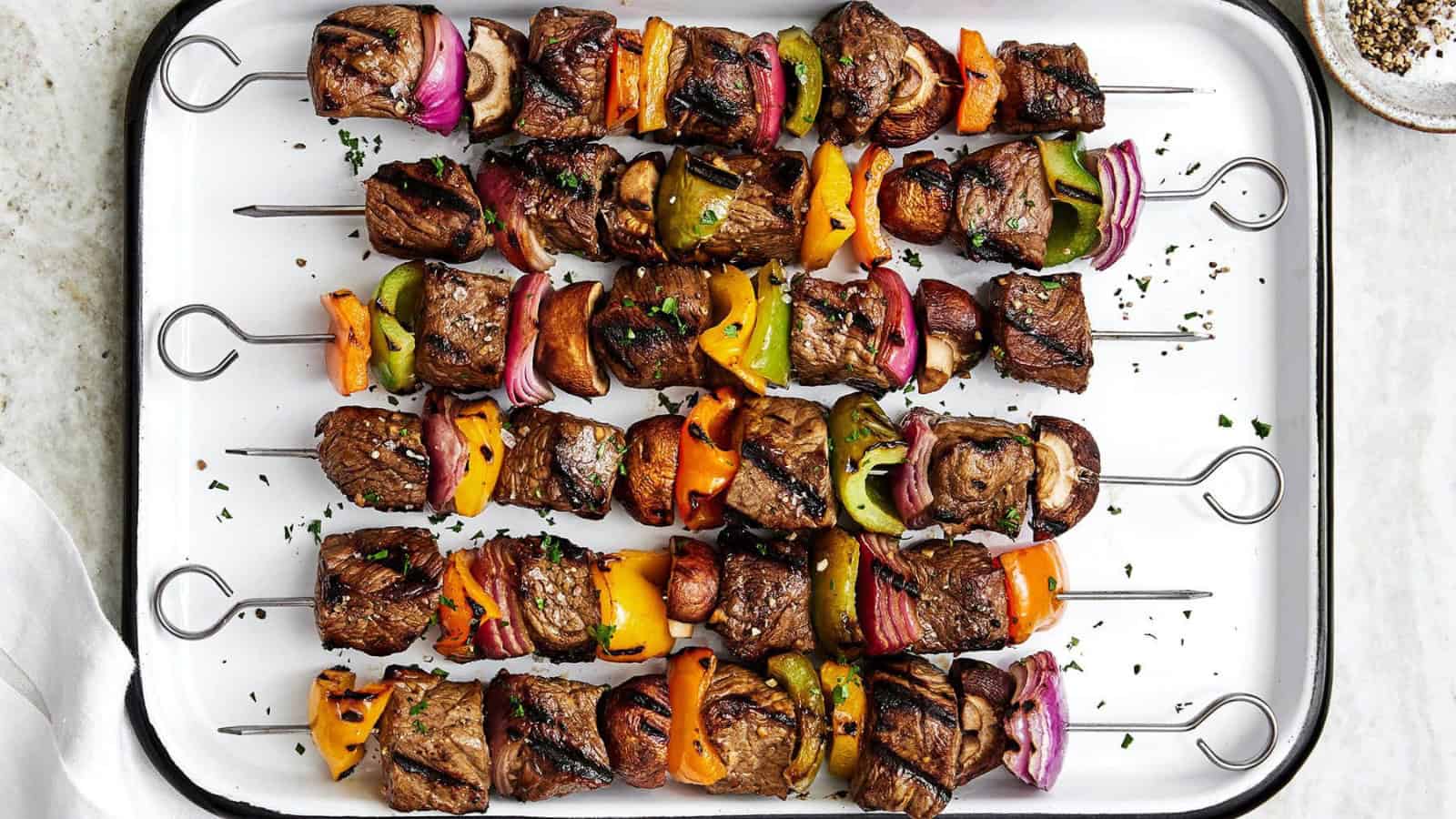 Skewers of grilled beef, mushrooms, bell peppers, and red onions arranged on a white platter. Garnished with fresh herbs.