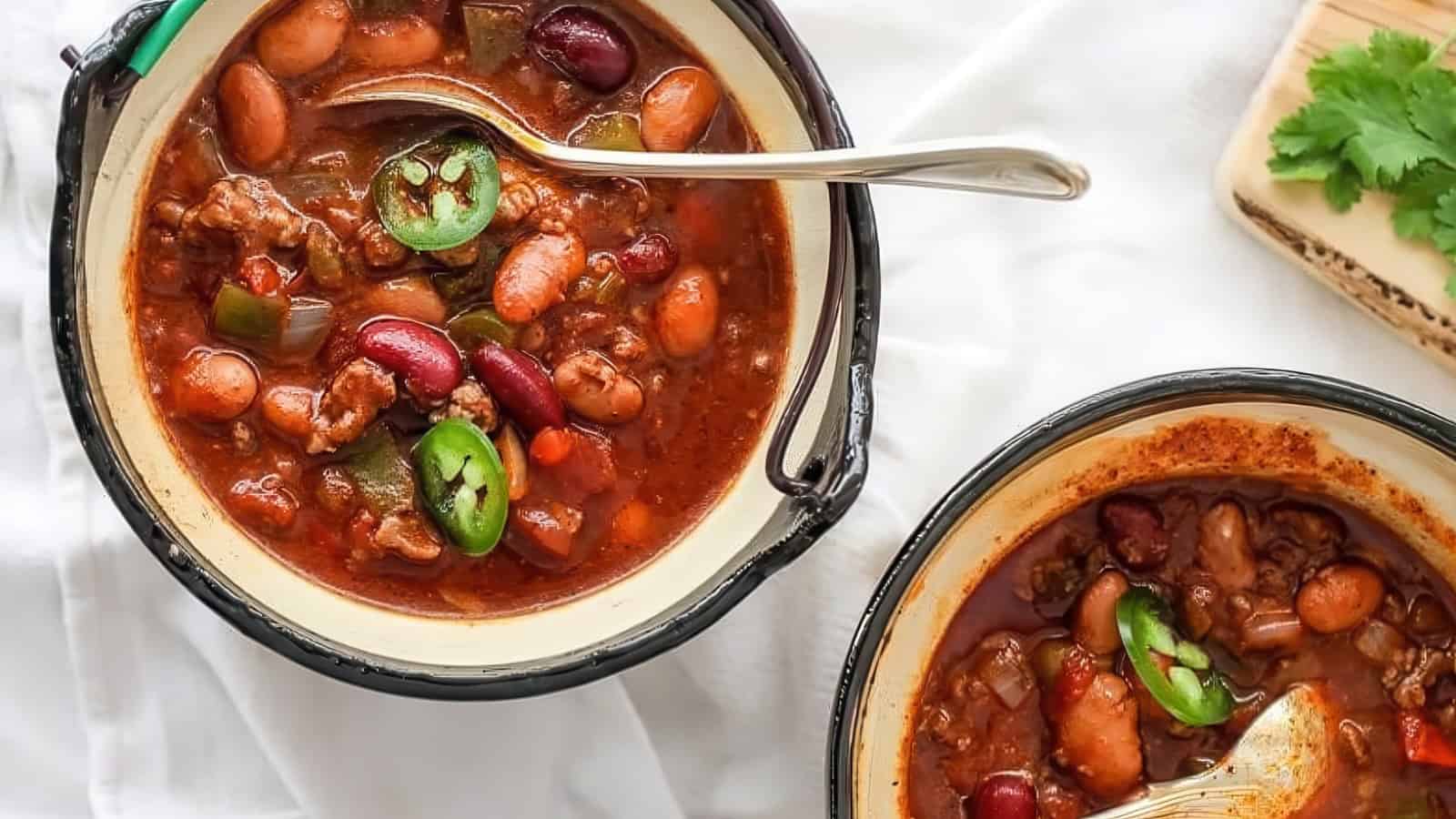Two bowls of chili filled with beans, ground meat, and bell peppers, garnished with sliced jalapeños. A spoon rests in one of the bowls. There is a sprig of cilantro beside the bowls. The background is a white cloth.