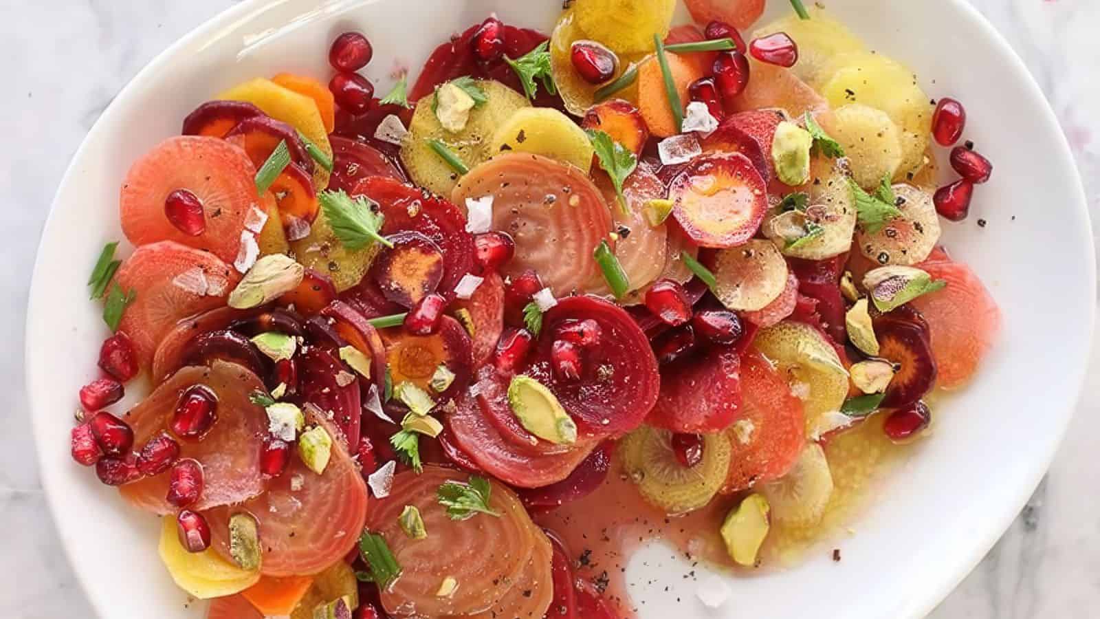 A colorful salad with thinly sliced rainbow carrots and beets, garnished with pomegranate seeds, pistachios, chopped herbs, and a sprinkling of seasoning on a white plate.