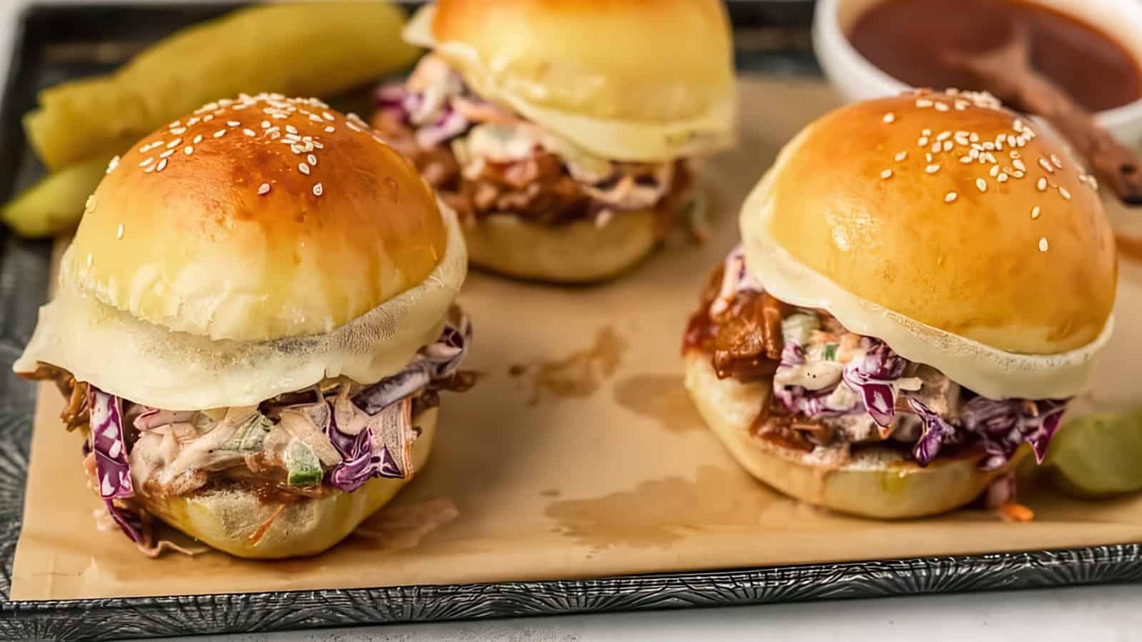 Three pulled pork sliders with coleslaw and melted cheese on sesame seed buns are arranged on a parchment-lined tray. A pickle and a bowl of barbecue sauce are in the background.
