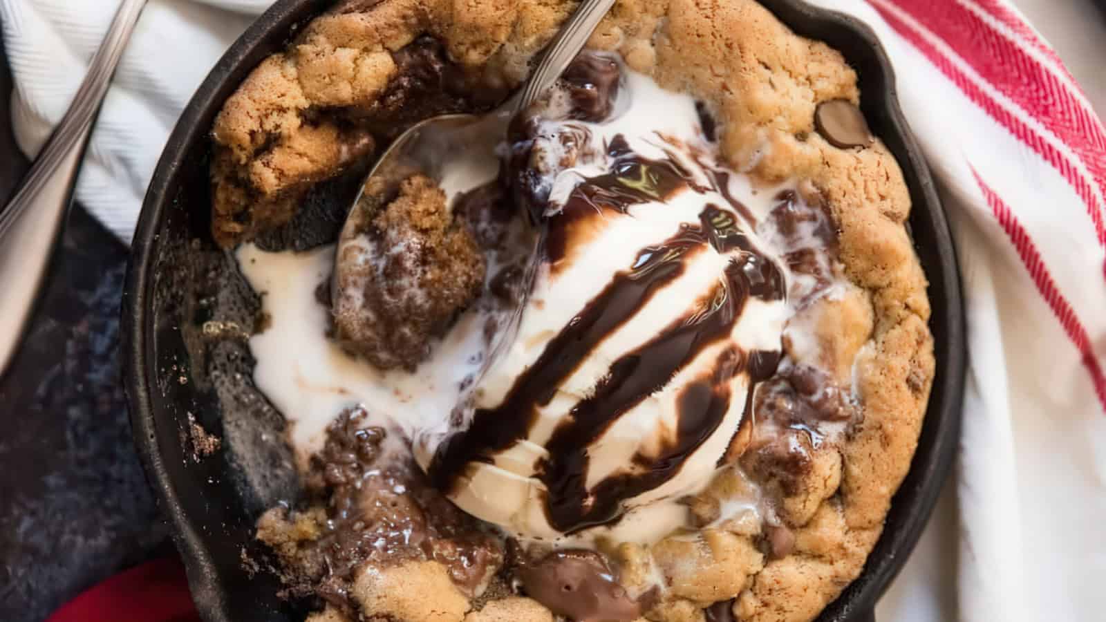 A warm, gooey chocolate chip cookie served in a skillet, topped with a scoop of melting vanilla ice cream and drizzled with chocolate sauce. A spoon rests in the dessert, and a red-striped towel is visible in the background.