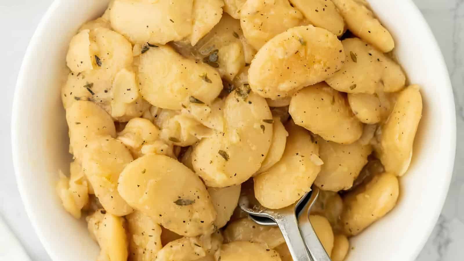 A bowl filled with creamy, cooked butter beans seasoned with herbs and black pepper. A spoon is resting in the bowl, ready to serve.