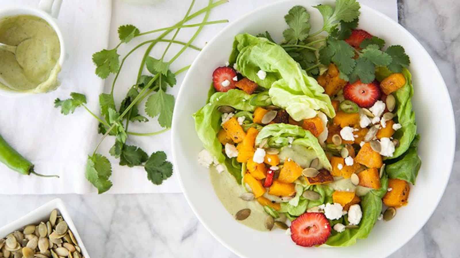 A fresh salad on a white plate featuring lettuce, roasted butternut squash, strawberries, pumpkin seeds, and crumbled cheese. Garnished with cilantro. Accompanied by a small bowl of dressing and cilantro sprigs on the side.