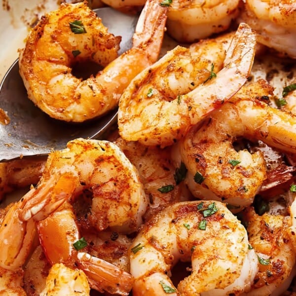 A dish of seasoned and cooked shrimp garnished with chopped herbs. The shrimp are orange and lightly browned, indicating they are well-cooked. A spoon rests among the shrimp in the pan.