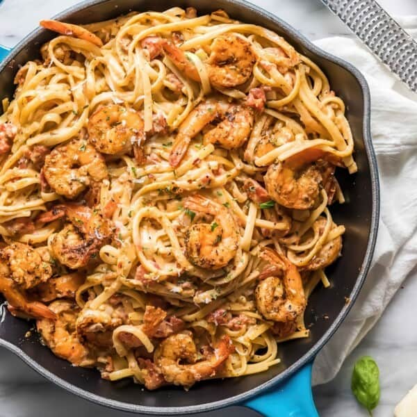 A skillet filled with creamy shrimp pasta garnished with herbs. The dish includes linguine, shrimp, and a creamy sauce. Surrounding the skillet are a cheese grater, wooden bowl with chili flakes, a block of cheese, and a pepper grinder.