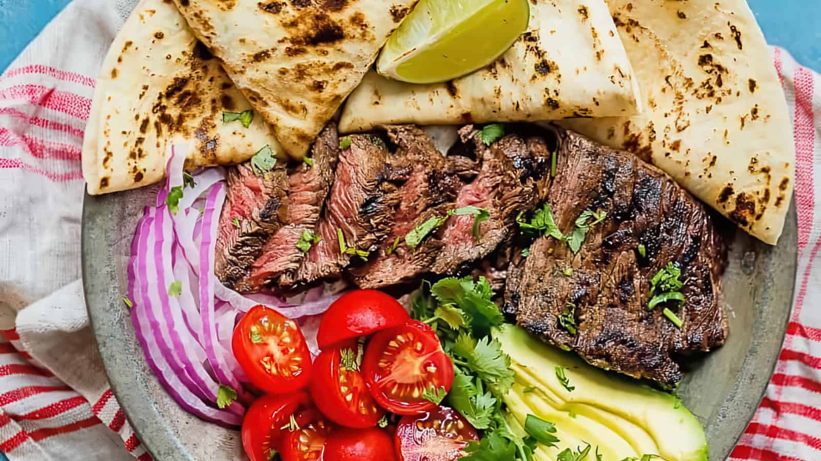 A plate with grilled beef slices, avocado, cherry tomatoes, red onion, and cilantro, garnished with lime wedges. Served with charred tortillas on the side, all atop a red-striped cloth. Served with