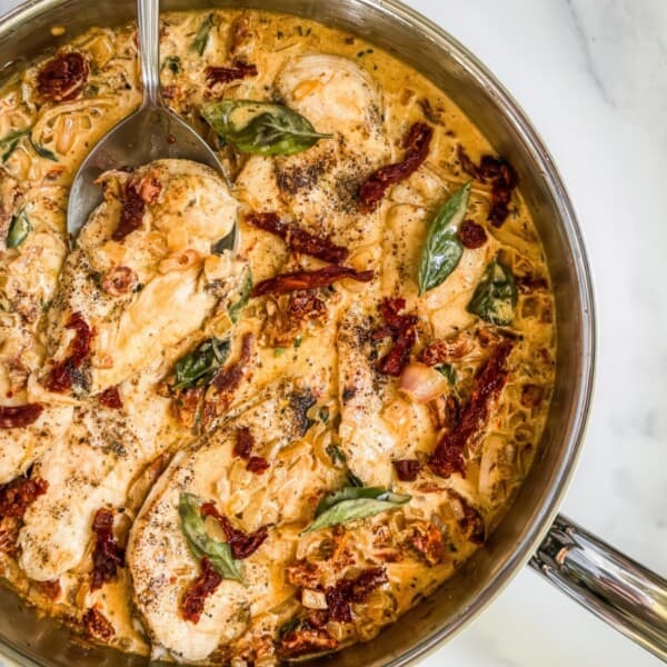 The image shows a creamy chicken dish in a skillet, featuring sun-dried tomatoes, basil, and pepper. It is presented on a marble surface.
