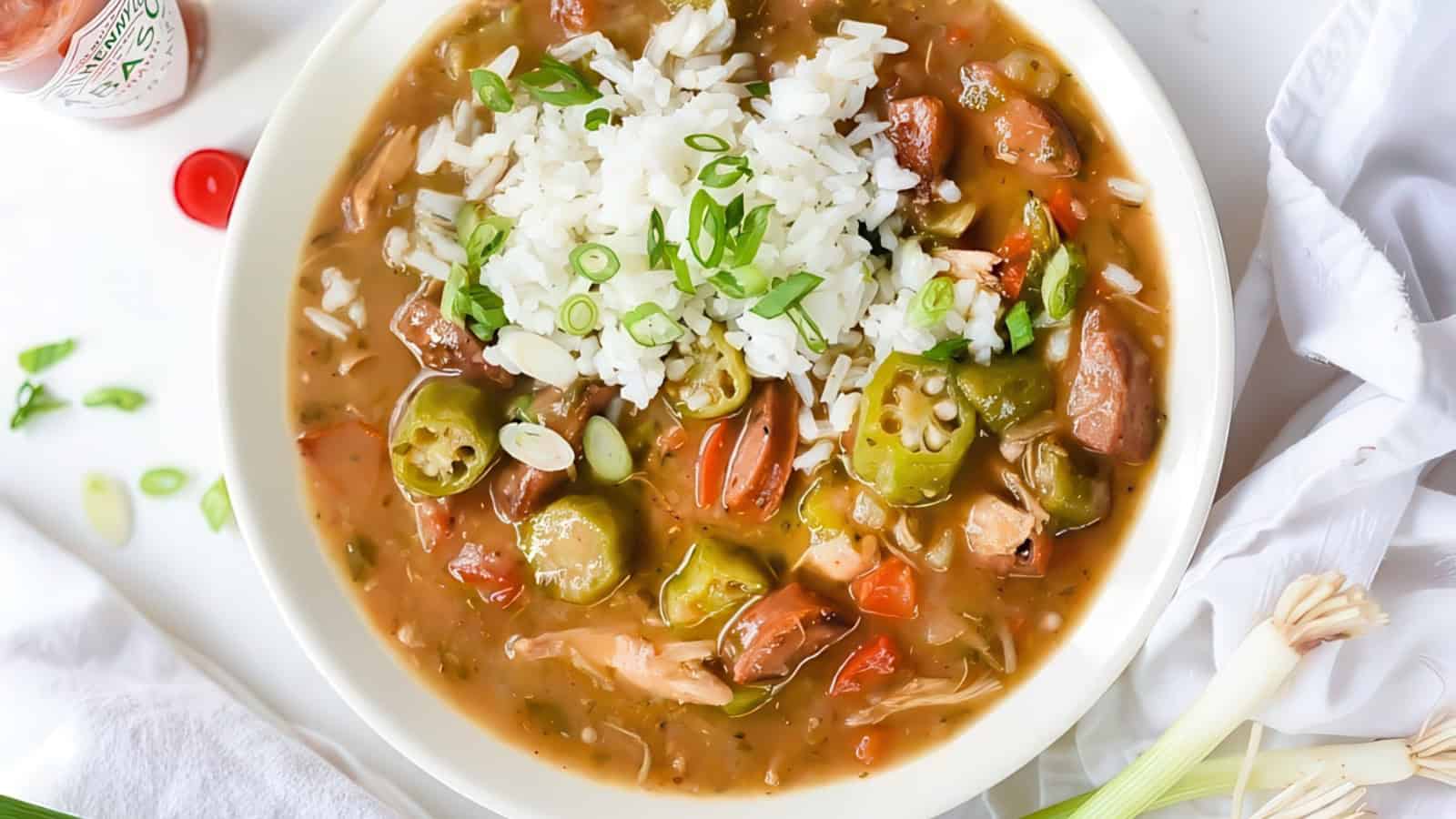 A bowl of gumbo topped with white rice and garnished with sliced green onions. The stew contains okra, sausage, chicken, and various vegetables in a rich, savory broth, placed on a white background with a hint of hot sauce nearby.