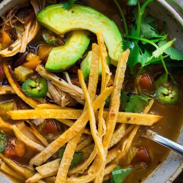A bowl of chicken tortilla soup topped with avocado slices, jalapeños, cilantro, and crispy tortilla strips. A spoon rests inside the bowl, sitting on a rustic wooden surface with a striped cloth nearby.