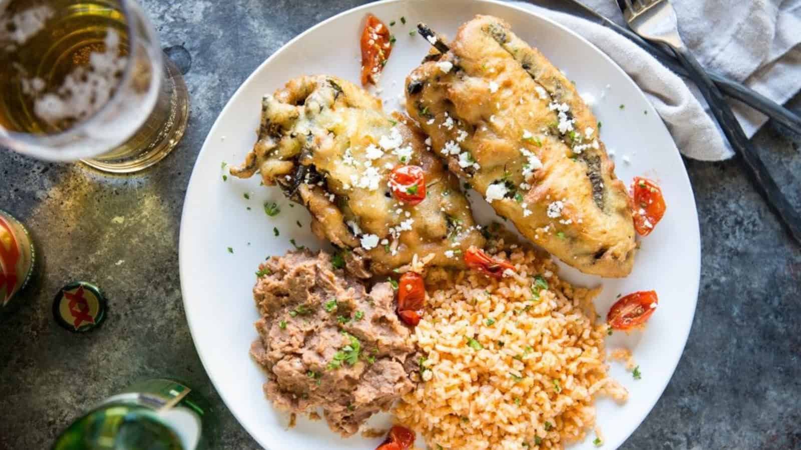 A plate with two chile rellenos topped with melted cheese, served alongside refried beans and Mexican rice. Garnished with chopped herbs and sliced cherry tomatoes. A glass of beer and utensils are nearby on a textured surface.