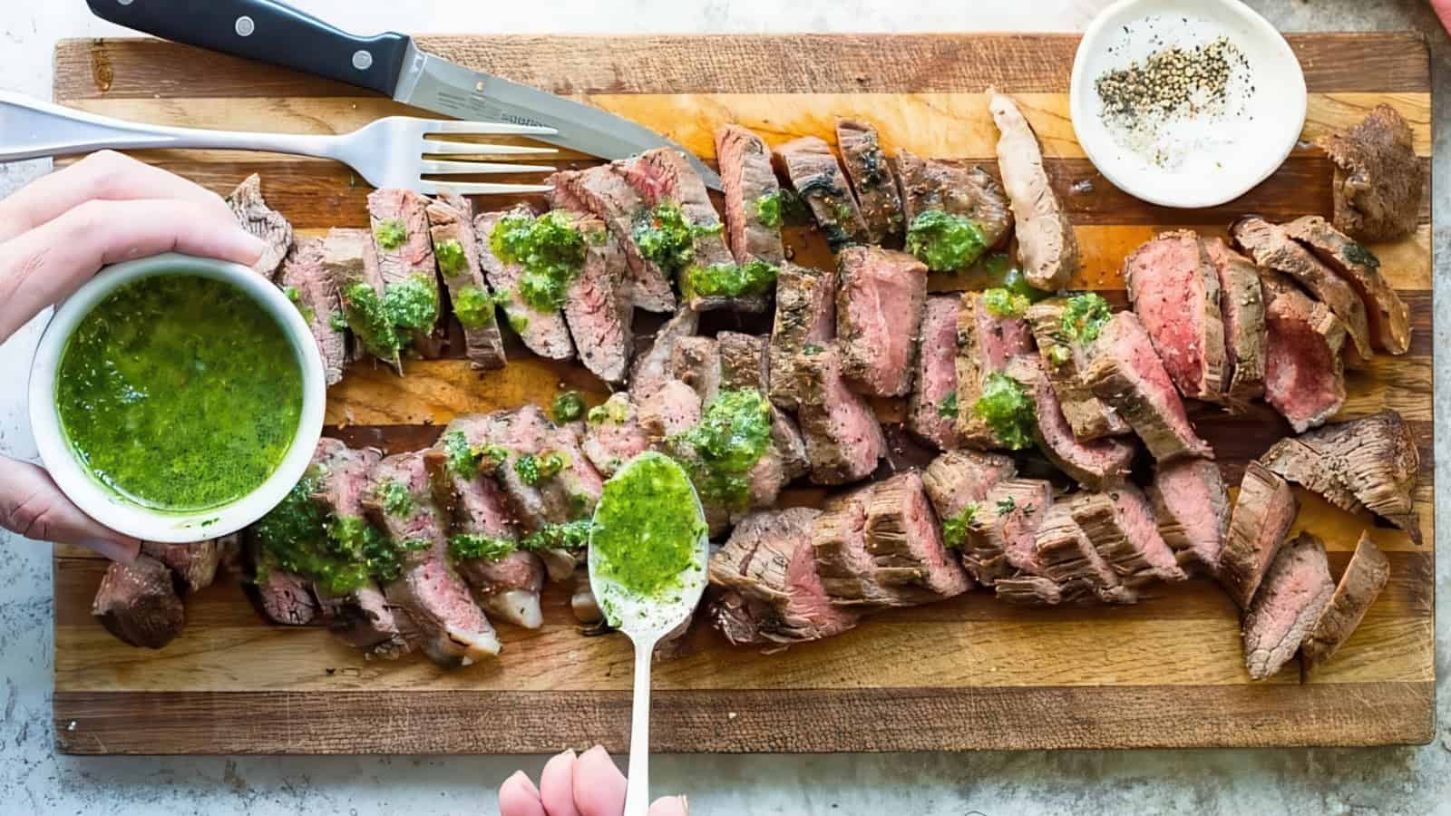 A wooden board with sliced grilled steak topped with green chimichurri sauce. A hand is pouring more sauce from a small bowl with a spoon. A fork, knife, and a small dish of pepper are nearby.