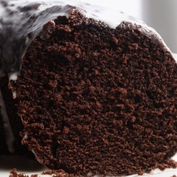 Close-up of a sliced chocolate loaf cake with a glossy white icing drizzled on top. The slice shows the cake's rich, moist texture. The background is softly blurred.