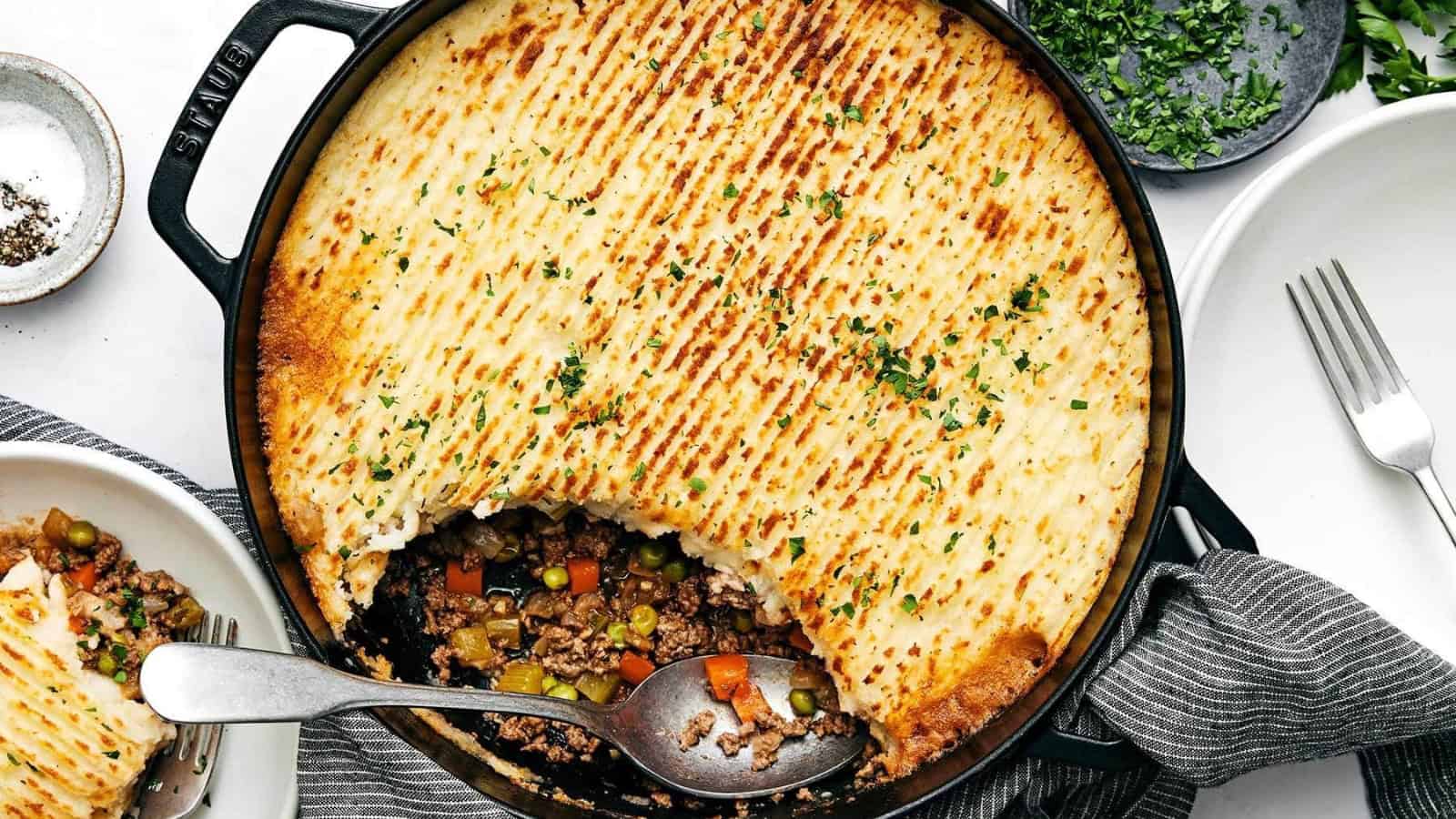A shepherd's pie in a skillet with a portion missing, revealing a layer of ground meat and vegetables beneath the baked mashed potato topping, garnished with herbs. A spoon rests inside, and a serving is on a small plate nearby. Utensils and napkin are also visible.