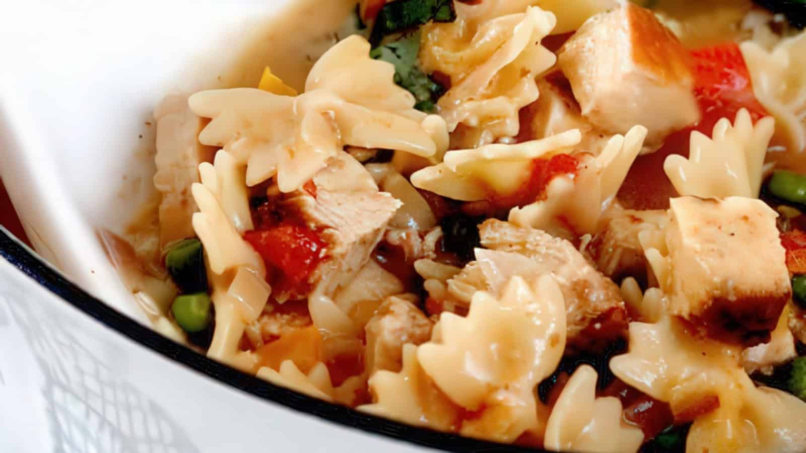 A close-up of bowtie pasta mixed with diced chicken, red bell peppers, green peas, and herbs, all in a creamy sauce. The dish is served in a white bowl.