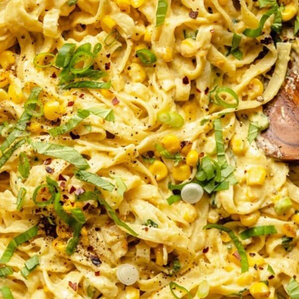 A skillet filled with creamy pasta topped with corn, chopped green onions, and black pepper. A wooden spoon rests on the side, revealing the rich texture of the sauce and garnishes.